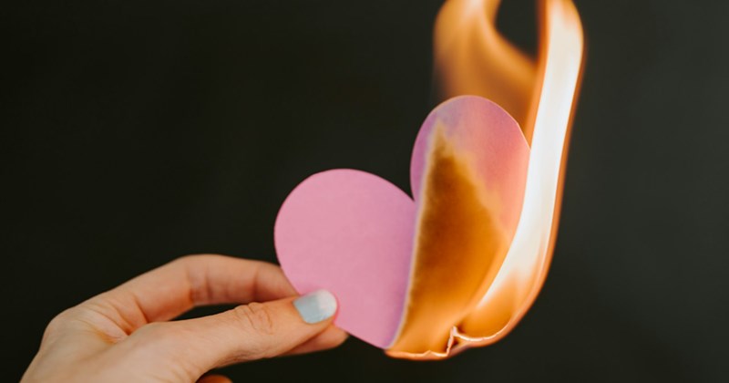 A woman's hand holding a paper heart on fire against a dark background.