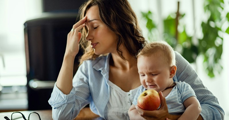 A mom sits down, holding a baby and an apple in one hand and rubbing her forehead with the other. 