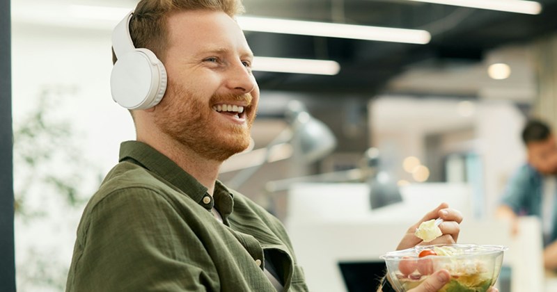 An employee wearing headphones smiles as he sits in an office eating salad.