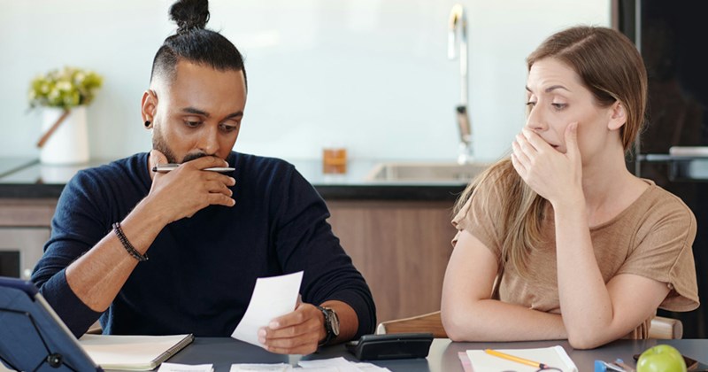 A young couple sitting at a table at home put their hands to their mouths as they look over financial papers. 
