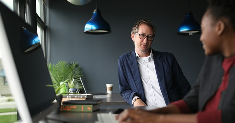 A male employee leans on a desk, speaking to a female coworker working on a computer in front of him. 
