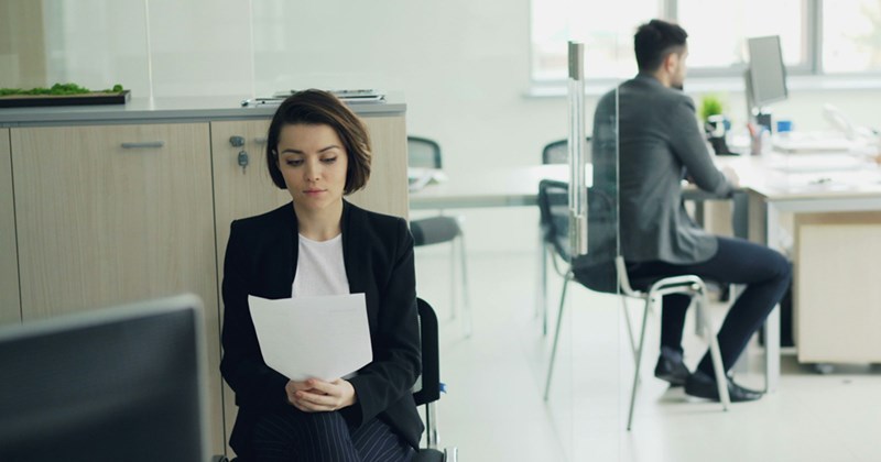 A woman sits outside a glass-walled office with two people in it, reading a piece of paper in her hand. 