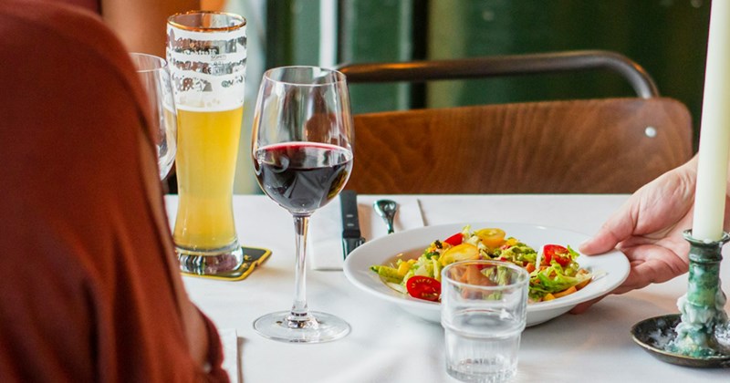 Close-up of a server putting a salad on a table that has several drinks glasses, and a couple sat there.