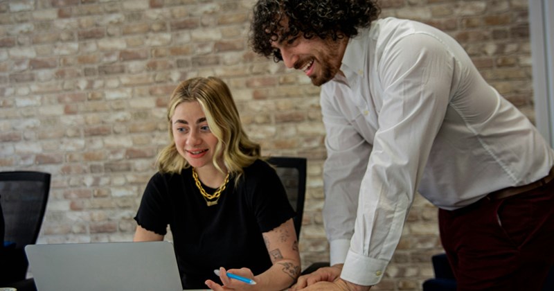 A male supervisor standing over a female employee working on a laptop.