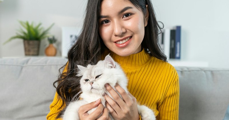A woman smiles while sitting on a couch and holding a white cat. 