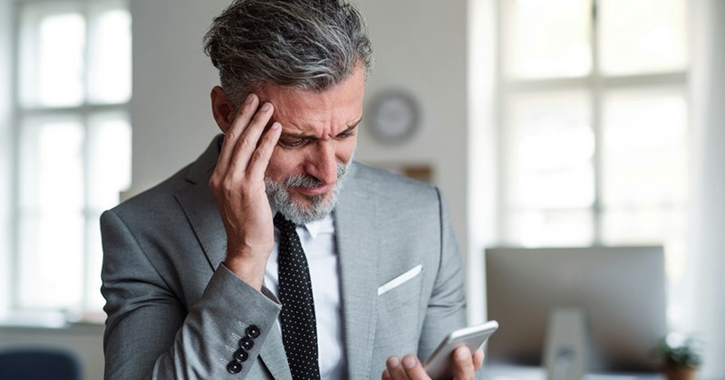 An older boss rubs his temple in frustration as he looks at his phone.