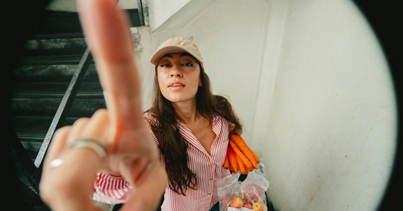 A woman holding grocery bags in one arm reaches a finger towards the fisheye lens of a camera.