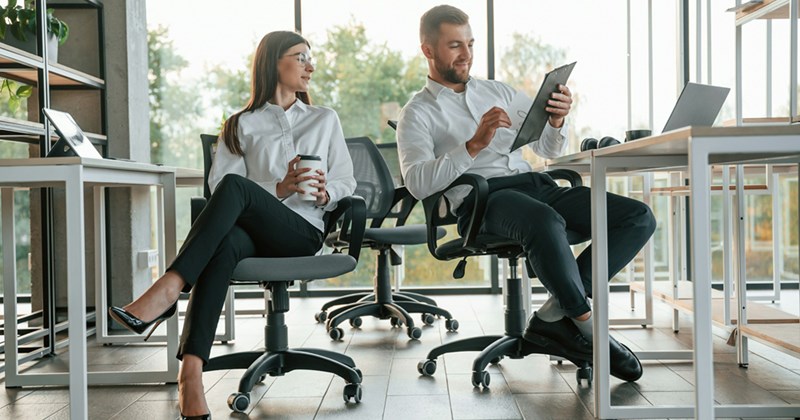 An employee and her boss work at opposite desks, with her pushing her chair towards him as he shows her something on a clipboard. 