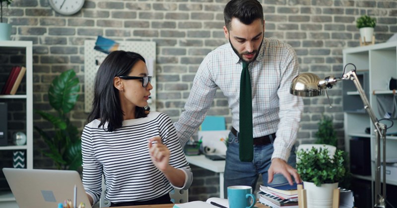 A male employee leans over a female colleague working at her desk, gesturing to some files on the desk.