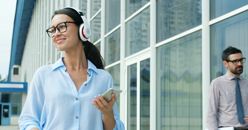 An office worker walks outside while holding her phone and listening to music on headphones.