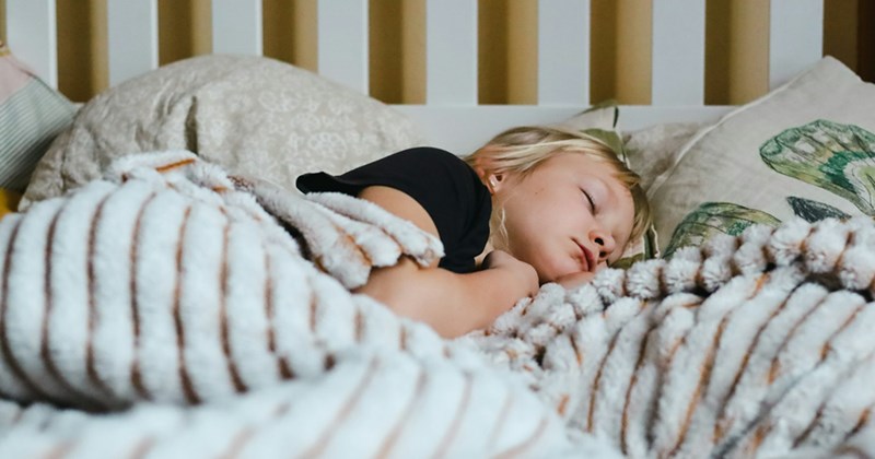 A young child sleeps on their side under a blanket in a white wooden frame bed. 