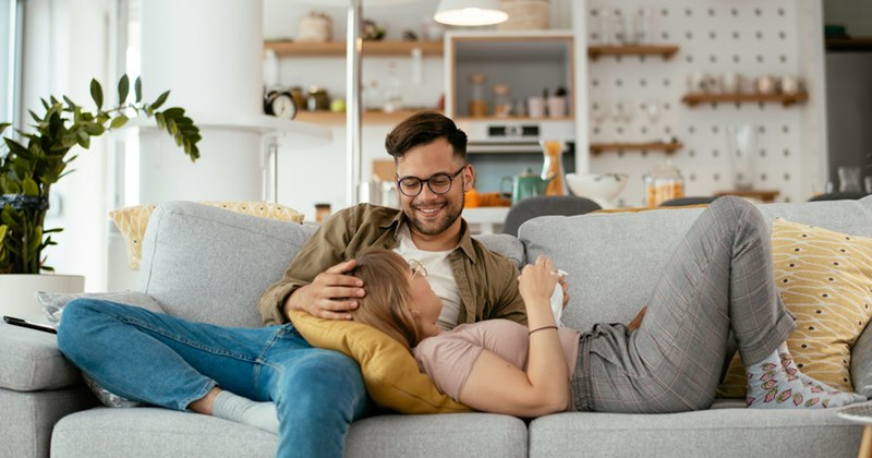 A couple relaxes on a couch, the woman resting in the man's lap with her head on a cushion.