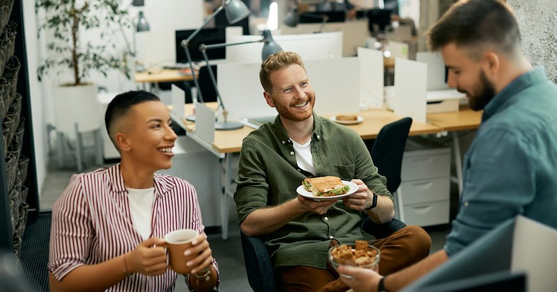 Three coworkers sit and eat lunch together with their open-plan office behind them.