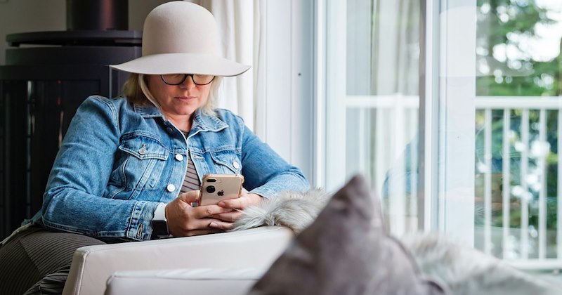 A woman with her face partially obscured by her hat sits by a window and uses her phone.