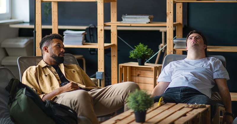 Two men sit around a coffee table, with one throwing his head back.