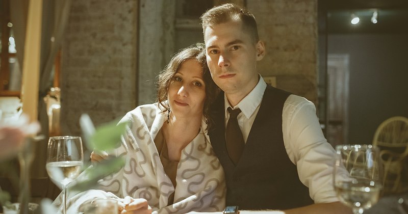 A woman leans on the shoulder of a man as they sit at a dining table in a restaurant. 