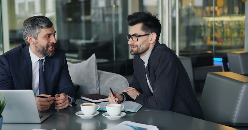 A boss and an employee sit at a table with coffee cups, a laptop, and notebooks, discussing something.