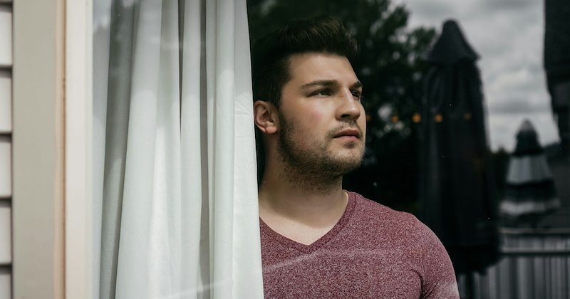 A man looks out a window while standing beside a curtain. 