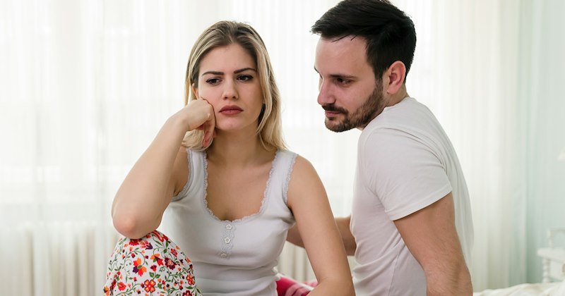 A woman sits on a bed looking upset, with a man beside her trying to console her.