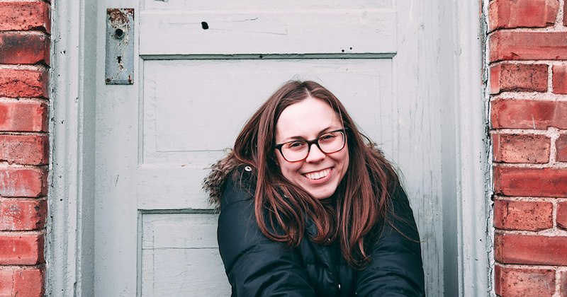 A woman wearing a coat smiles as she sits in front of a door on a brick wall. 