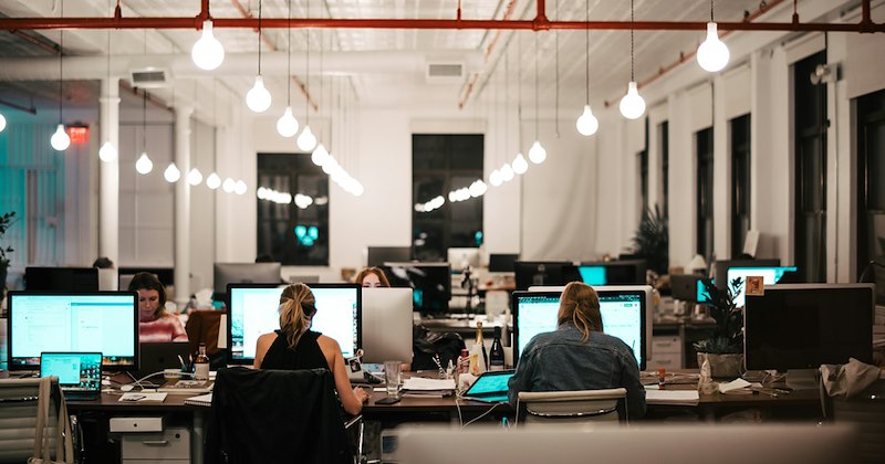Several employees working at computers in an open-plan office with hanging lights.