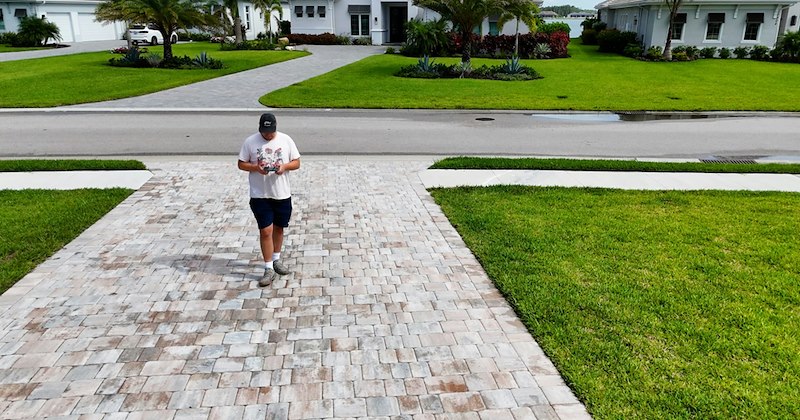 A man walks up a wide driveway in a suburban neighborhood while looking at his phone.