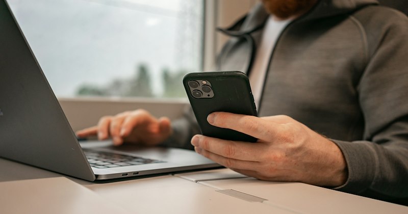 Close-up of a man sitting at a laptop while holding his phone.