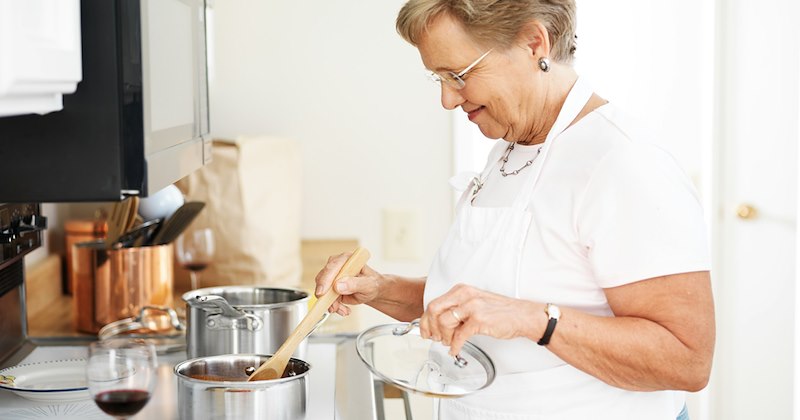 A grandma holds a pan lid in one hand and stirs a pot on a stove with a wooden spoon in the other. 