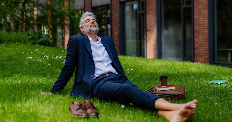 A businessman relaxes barefoot on the grass, his shoes and briefcase on either side of him.