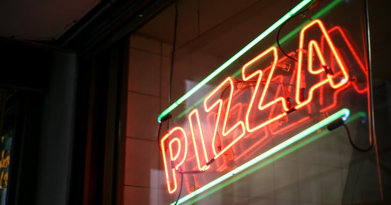 A red and green neon sign in a restaurant window that says ‘PIZZA’. 