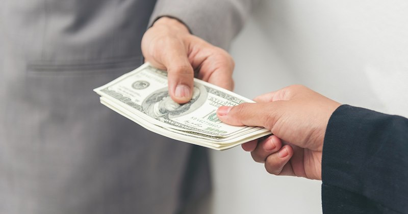 Close-up of the arm of one person in business attire handing another person a stack of dollar bills. 
