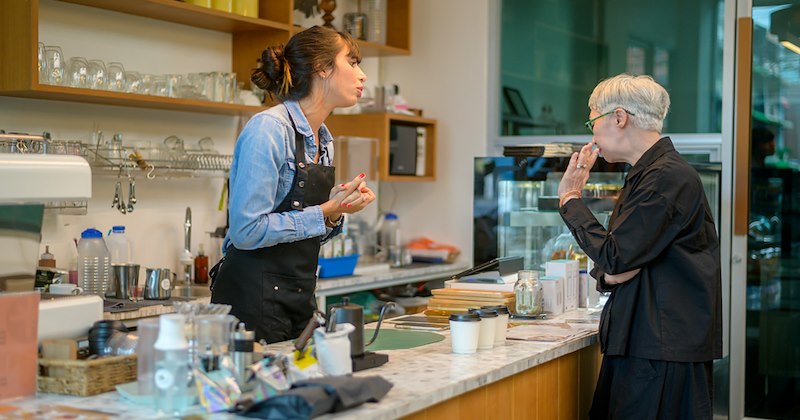 An angry older woman waves her hand at the barista behind the counter who is talking to her in a coffee shop.