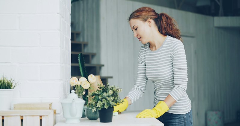 A woman cleans a table with flowers and knick-knacks on it while wearing yellow gloves.