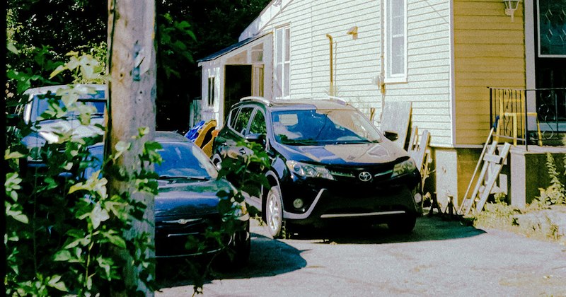 Two cars parked in a driveway in front of the porch of a yellow house. 