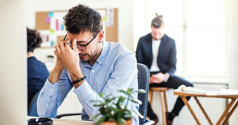 An employee pinches the bridge of his nose while taking a phone call at his desk, with colleagues sitting in the background. 